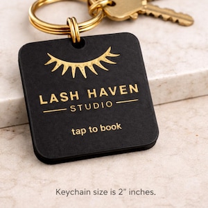 Może przedstawiać: Czarny kwadratowy brelok do kluczy ze złotym tekstem i złotym wzorem rzęs. Tekst brzmi "LASH HAVEN STUDIO" i "tap to book". Brelok jest przymocowany do złotego kółka i dwóch złotych kluczy. Rozmiar: 5 cm.
