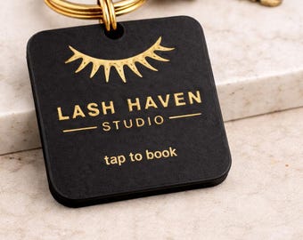 Puede incluir: Llavero cuadrado negro con texto dorado y diseño de pestañas doradas. El texto dice "LASH HAVEN STUDIO" y "tap to book". El llavero está unido a un llavero dorado y dos llaves doradas. Tamaño: 5 cm.