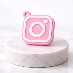 May include: A pink Instagram camera charm with white detailing. The charm, square in shape, features a circular lens and a small loop. It rests on a white marble pedestal.