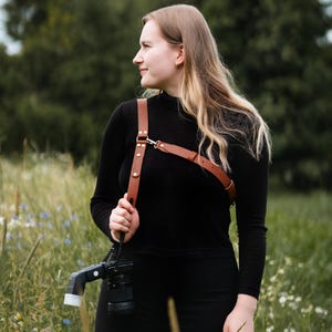 May include: A woman wearing a black top and black trousers with a brown leather camera strap. She is holding a camera with a flash attached to it. The woman is standing in a field of green grass and wildflowers.