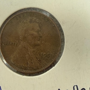 Puede incluir: Una moneda de cobre de 1951 de Lincoln, con un perfil de Abraham Lincoln. La moneda está dentro de un soporte de plástico transparente. La palabra "LIBERTY" es visible en la moneda.