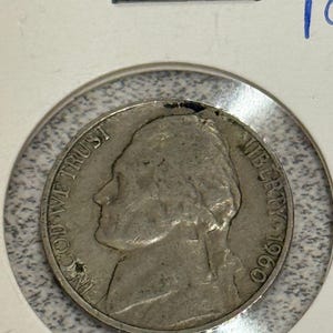 May include: A silver 1960 Jefferson nickel coin, showing a profile of Thomas Jefferson and the words "In God We Trust" and "Liberty". The coin is in a protective holder with a blue number written on the top right.