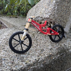May include: A red and black toy mountain bike with a wooden frame and black wheels. The bike is sitting on a gray concrete surface.