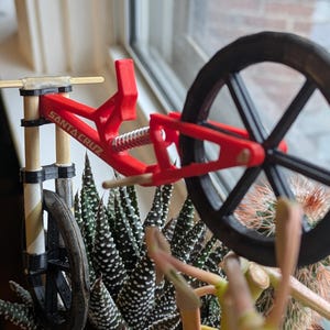 May include: A red and black miniature bicycle with a wooden frame and a black wheel. The bicycle has a spring suspension and the words "SANTA CRUZ" printed on the frame.