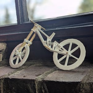Puede incluir: Un modelo blanco de una bicicleta de monta&ntilde;a con una horquilla de suspensi&oacute;n y un cuadro blanco. La bicicleta est&aacute; sentada sobre una superficie de ladrillo.