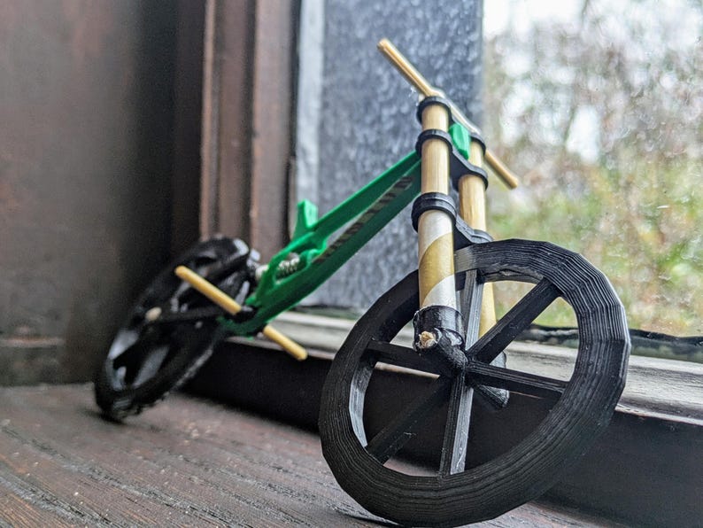 Puede incluir: Una bicicleta verde y negra en miniatura con manillar y ruedas de madera. La bicicleta est&aacute; sobre una superficie de madera.