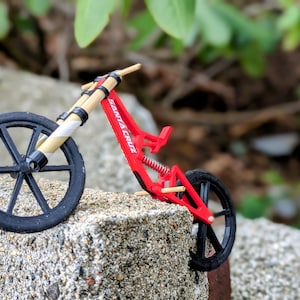 May include: A red and black miniature bicycle with a wooden handlebar and the words "Santa Cruz" printed on the frame. The bicycle is sitting on a gray rock.