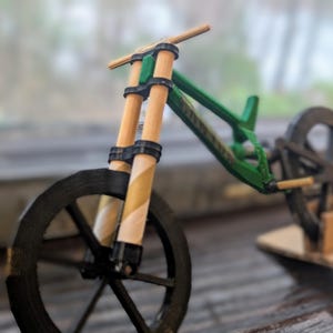 Puede incluir: Una bicicleta de monta&ntilde;a en miniatura verde y negra con manillar de madera y ruedas negras. La bicicleta est&aacute; sobre un soporte de cart&oacute;n.