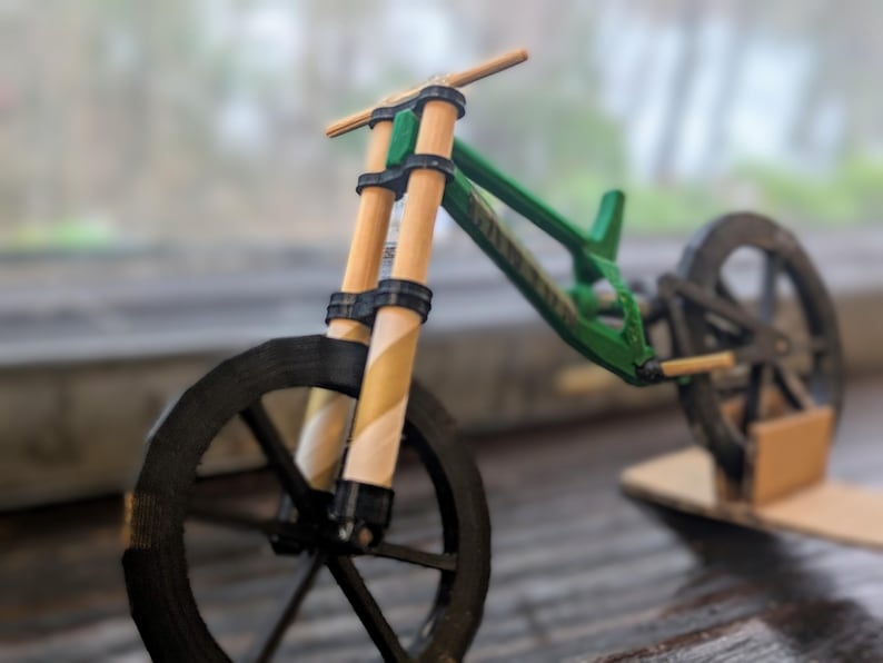 Puede incluir: Una bicicleta de monta&ntilde;a en miniatura verde y negra con manillar de madera y ruedas negras. La bicicleta est&aacute; sobre un soporte de cart&oacute;n.
