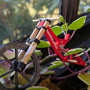 Könnte beinhalten: Ein rotes und schwarzes Miniatur-Mountainbike mit dem Schriftzug "Santa Cruz" auf dem Rahmen. Das Fahrrad ist aus Kunststoff und hat einen Holzlenker.