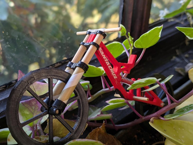 May include: A red and black miniature mountain bike with the words "Santa Cruz" printed on the frame. The bike is made of plastic and has a wooden handlebar.