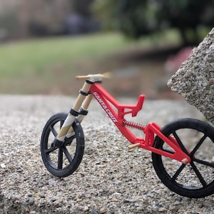 May include: A red and black miniature mountain bike with a suspension fork and the word "Santa Cruz" printed on the frame.