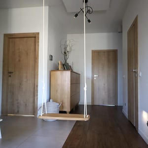 Op de afbeelding: Een houten schommel hangt aan witte touwen aan het plafond. Het interieur heeft houten deuren, een houten dressoir en een donkere houten vloer. De muren zijn wit geschilderd. De kamer heeft een modern design.