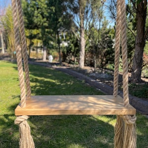 May include: A wooden swing set with natural rope hanging from a tree branch. The swing is made of a single piece of wood and has a natural finish. The rope is thick and has a natural colour.