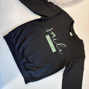 Peut inclure: Sweat-shirt noir à manches longues et col rond. Le sweat-shirt porte l'inscription "smile EVERY DAY" en vert clair. Les poignets et l'ourlet sont côtelés.