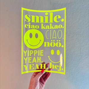 Könnte beinhalten: Ein transparenter neongelber Aufkleber mit den Worten "smile," "ciao kakao," "yippie yeah," und "yeah hej." Ein Smiley-Gesicht ist ebenfalls enthalten. Der Aufkleber wird von einer Person gehalten.
