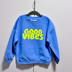 Può includere: Una felpa blu con la scritta "GOOD VIBES" in giallo brillante, appesa a una gruccia nera. La felpa ha un girocollo e maniche lunghe. Lo sfondo è un muro bianco.