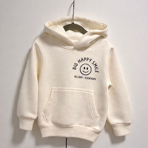Könnte beinhalten: Cremefarbener Kapuzenpullover mit Fronttasche und Kapuze. Auf der Vorderseite befindet sich eine schwarze Grafik eines lächelnden Gesichts mit den Worten "BIG HAPPY SMILE ALL DAY - EVERYDAY". Der Hoodie hängt an einem Kleiderbügel.