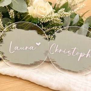 Può includere: Due segnaposto rotondi in acrilico trasparente con i nomi "Laura" e "Christoph" in corsivo rosa, ciascuno con un motivo a pennellate verde salvia e un piccolo cuore. I biglietti sono appoggiati su un tessuto bianco con composizioni floreali.