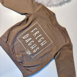 Könnte beinhalten: Ein braunes Sweatshirt mit dem Text "FRESH DACHS" in einem weißen Quadrat und dem Namen "LIAM" darunter. Das Sweatshirt hat lange Ärmel und gerippte Bündchen und Saum. Im Hintergrund befindet sich ein flauschiger weißer Gegenstand.