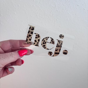 Könnte beinhalten: Transparenter Aufkleber mit dem Wort "hej." in schwarzen Buchstaben mit Leopardenmuster. Der Aufkleber wird vor einer weißen Wand gehalten.