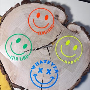 Könnte beinhalten: Vier bunte Smiley-Aufkleber auf einer Holzoberfläche. Die Aufkleber sagen "Kita Kind", "Schulkind", "Happy" und "Whatever" mit einem Kreuzsymbol.