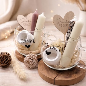 Könnte beinhalten: Zwei klare Glastassen mit weißen und malvenfarbenen Stabkerzen, weißen runden Kerzenhaltern und herzförmigen Holzsteckern. Die Tassen sind mit dem schwarzen Schriftzug DANKE und FUR DICH sowie Herzsymbolen versehen und stehen auf Holzuntersetzern.