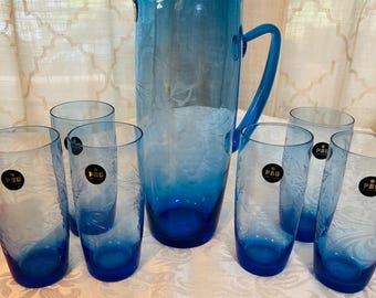 May include: A blue glass pitcher and matching drinking glasses. The pitcher has a handle and a floral design. The glasses are cylindrical and have a dark blue base. The set is arranged on a white tablecloth.