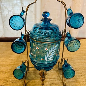 May include: A blue glass liquor dispenser with a metal stand and six matching shot glasses. The dispenser has a white floral design and a spigot.