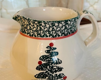 May include: Cream-colored ceramic pitcher with a green speckled rim and a red stripe. The pitcher features a Christmas tree design with red ornaments. It has a handle and a spout.