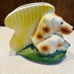 May include: A ceramic dog planter featuring two dachshunds with brown spots, cream-colored fur, and black noses. The planter has a yellow, fan-shaped background and a green base. A decorative item for dog lovers.