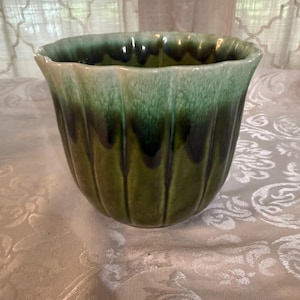 May include: A green ceramic planter with a fluted design. The planter has a glossy finish with a gradient of green hues, transitioning from a lighter shade at the top to a darker shade at the bottom. The rim has a wavy edge.