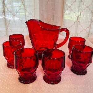 May include: A red glass pitcher and six matching tumblers. The pitcher has a handle and a spout. The tumblers are short and wide. All pieces have a textured, geometric design. The set is arranged on a white surface.