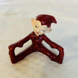 May include: A red ceramic figurine of an elf sitting with its legs crossed. The elf has a white face with painted features and a red hat.