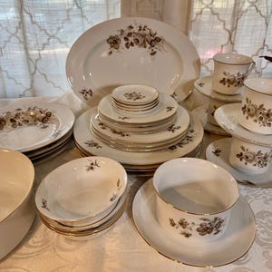 May include: A collection of white porcelain dinnerware with a floral design in shades of brown and gold trim. The set includes plates, bowls, cups, saucers, and a serving platter, perfect for a formal dining experience.