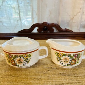 May include: A vintage cream-colored sugar bowl and creamer set with a floral pattern in shades of brown, yellow, and white. The set has a brown trim around the top.