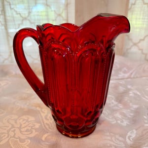May include: A ruby red glass pitcher with a handle and a spout. The pitcher has a decorative pattern of vertical lines and rounded shapes. The pitcher is sitting on a white surface.