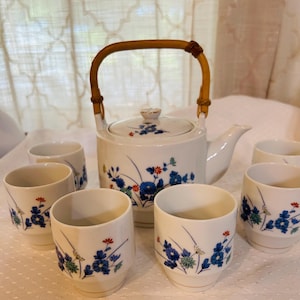 May include: A white ceramic tea set with a teapot and six cups. The teapot has a bamboo handle and is decorated with blue floral patterns. The cups also feature the same blue floral design with red and green accents.