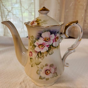 May include: A white porcelain teapot with a floral design. The teapot has a gold trim and a lid with a gold knob. The flowers are pink, purple, and green.