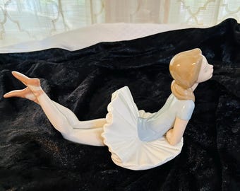 May include: A porcelain figurine of a ballerina in a white tutu and a light blue leotard. She is lying on her side with her head turned to the side and her eyes closed. Her hair is brown and pulled back in a ponytail.