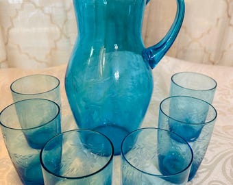 May include: A turquoise glass pitcher with a handle and six matching tumblers. The pitcher and glasses feature etched floral designs. The glassware set is ideal for serving beverages and adds a touch of elegance to any table setting.