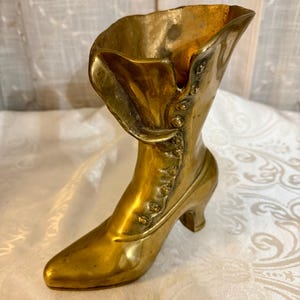May include: A vintage brass boot-shaped vase. The boot has a pointed toe, a small heel, and a folded-over top. Decorative buttons run down the side. The brass has a warm, aged patina, suggesting it is an antique decorative item.