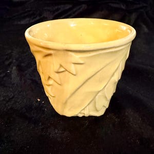 May include: A pale yellow ceramic planter featuring a raised, embossed leaf pattern across its exterior surface. The vessel has a wide, circular opening with a smooth rim and a tapered base, showcasing a glossy finish that reflects light.