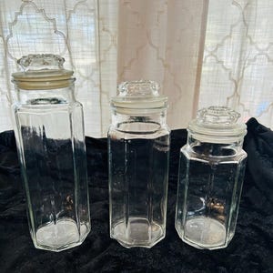May include: Three clear glass jars with lids. The jars are octagonal in shape and have a textured surface. The lids are made of glass and have a raised design.