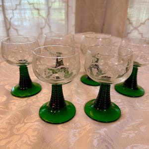 May include: Six clear wine glasses with etched floral designs. The stems are black with a green base. The glasses are arranged on a white patterned tablecloth. The glasses are vintage and elegant.