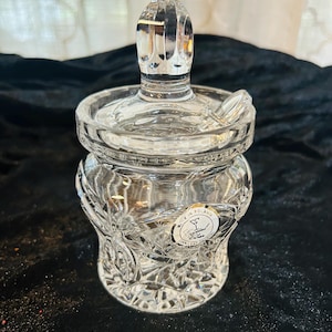May include: A clear, cut-glass jar with a lid and a decorative handle. The jar has a round body with a textured, floral-like pattern. The lid has a small notch for a spoon. The jar is labeled "Made in Poland".