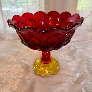 May include: A ruby red glass pedestal bowl with a scalloped rim and a yellow base. The bowl features a moon and star pattern. The stem is red. The bowl is approximately 8 inches in diameter and 6 inches tall.
