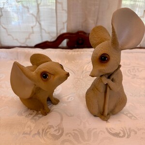 May include: Two tan mouse figurines with large ears and dark brown eyes. One mouse is in a seated position looking to the side, while the other mouse is standing upright and holding a thin, vertical rod-like object in its front paws.