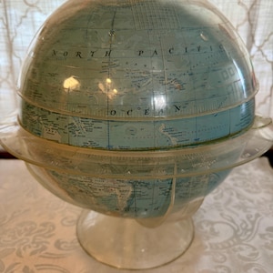 May include: A vintage globe with a clear plastic meridian and base. The globe features a light blue and white color scheme, with geographical details and text. The globe is a decorative item.