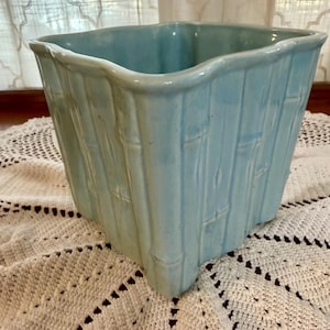 May include: A square, light blue ceramic planter with a bamboo-style texture. The planter has a slightly wavy rim and is approximately 8 inches wide and 7 inches tall. The planter is sitting on a white, crocheted doily.